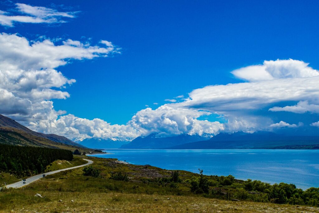 Blue sky with clouds, grass field, road, beautiful lake with mountains in the distance